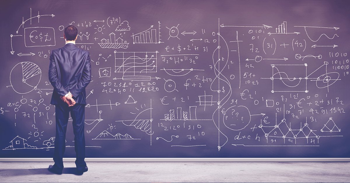 A man in a suit stands before a blackboard covered in charts, equations, and figures