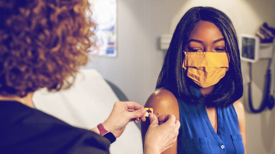 A nurse places a bandage on the upper arm of woman wearing a mask, presumably after giving her a vaccine
