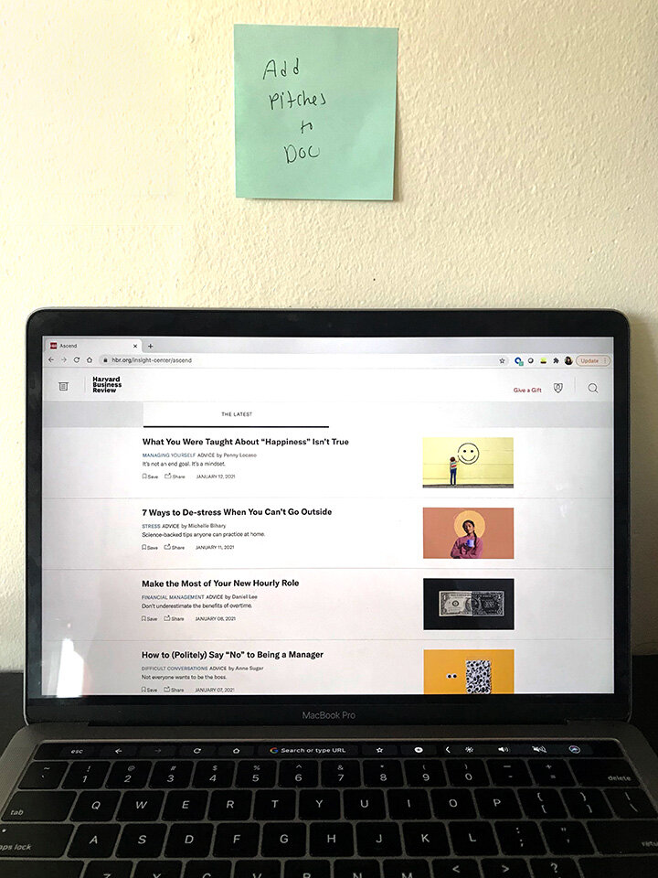 A laptop sits open in front of a wall bearing a sticky note that reads "Add Pitches to Doc"