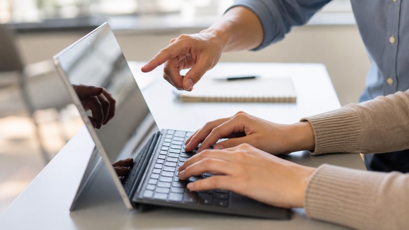 Workplace Collaboration at a Laptop Two business professionals collaborate at a laptop in a bright office. One person types while the other points at the screen, illustrating hands-on coaching, training, or collaborative problem-solving.